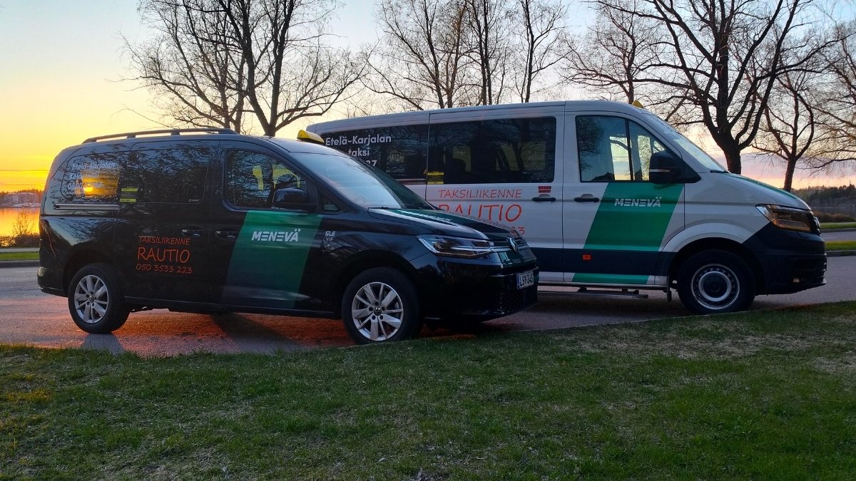 Kaksi taksiyrityksen pakettiautoa pysäköitynä tien viereen ilta-auringon laskiessa taustalla; vasemmalla musta pienempi ajoneuvo ja oikealla valkoinen suurempi pakettiauto, molemmissa vihreät ja punaiset teippaukset, joissa lukee muun muassa "RAUTIO" ja "MENEVÄ".