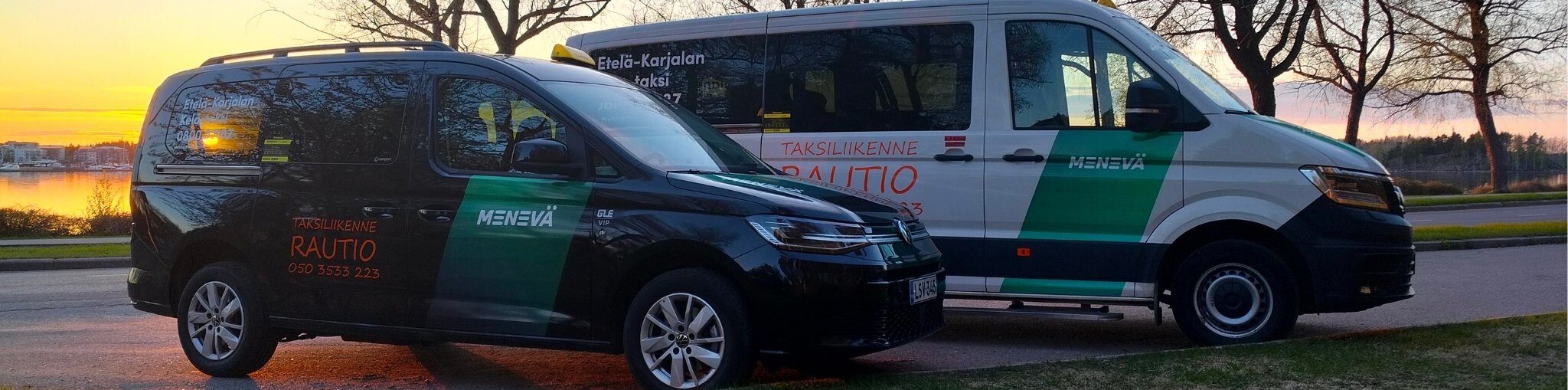 Kaksi taksiyrityksen pakettiautoa pysäköitynä tien viereen ilta-auringon laskiessa taustalla; vasemmalla musta pienempi ajoneuvo ja oikealla valkoinen suurempi pakettiauto, molemmissa vihreät ja punaiset teippaukset, joissa lukee muun muassa "RAUTIO" ja "MENEVÄ".